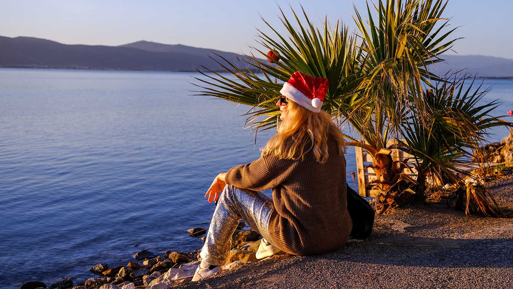 Woman in a Santa hat, sitting on a beach with palm leaves behind her, mountains in the distance.